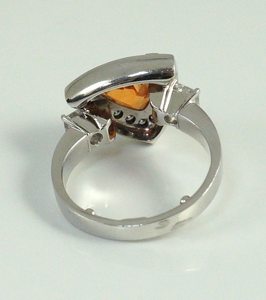 A modern platinum and single stone trilliant cut spessartite garnet ring, with diamond set border and two criss-emerald cut diamond set shoulders, by Musson
